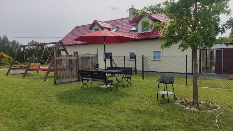 widok na huśtawki ogrodowe, duży parasol, pod nim stół i ławki, nieduże drzewo z boku, w tle jasny dom